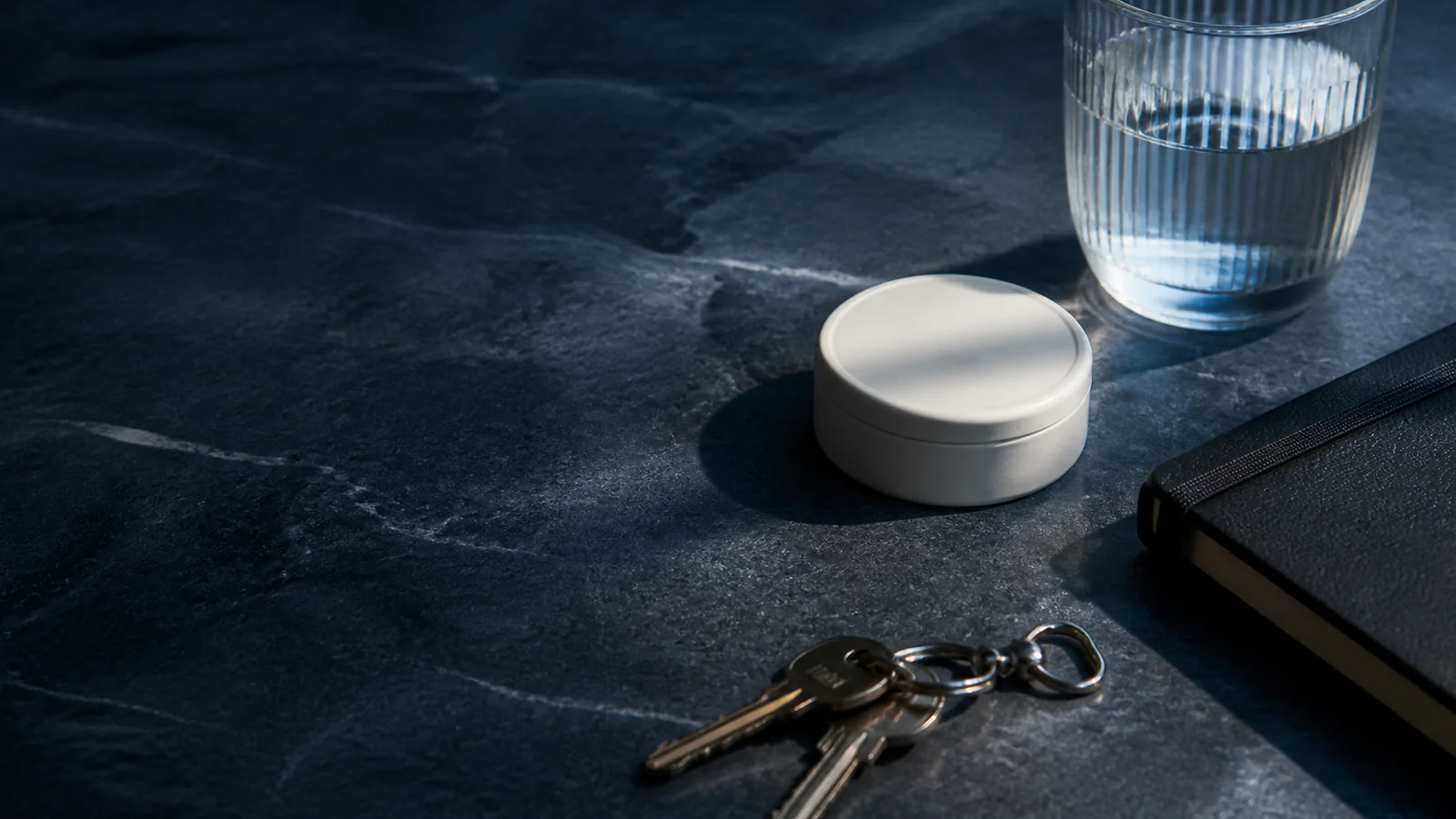 Unbranded round tin beside water, keys, and a notebook on a dark stone surface